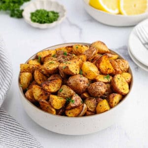A bowl filled with golden, roasted Air Fryer Potatoes, garnished with chopped herbs. In the background, there are lemon slices, parsley, and a plate with forks.