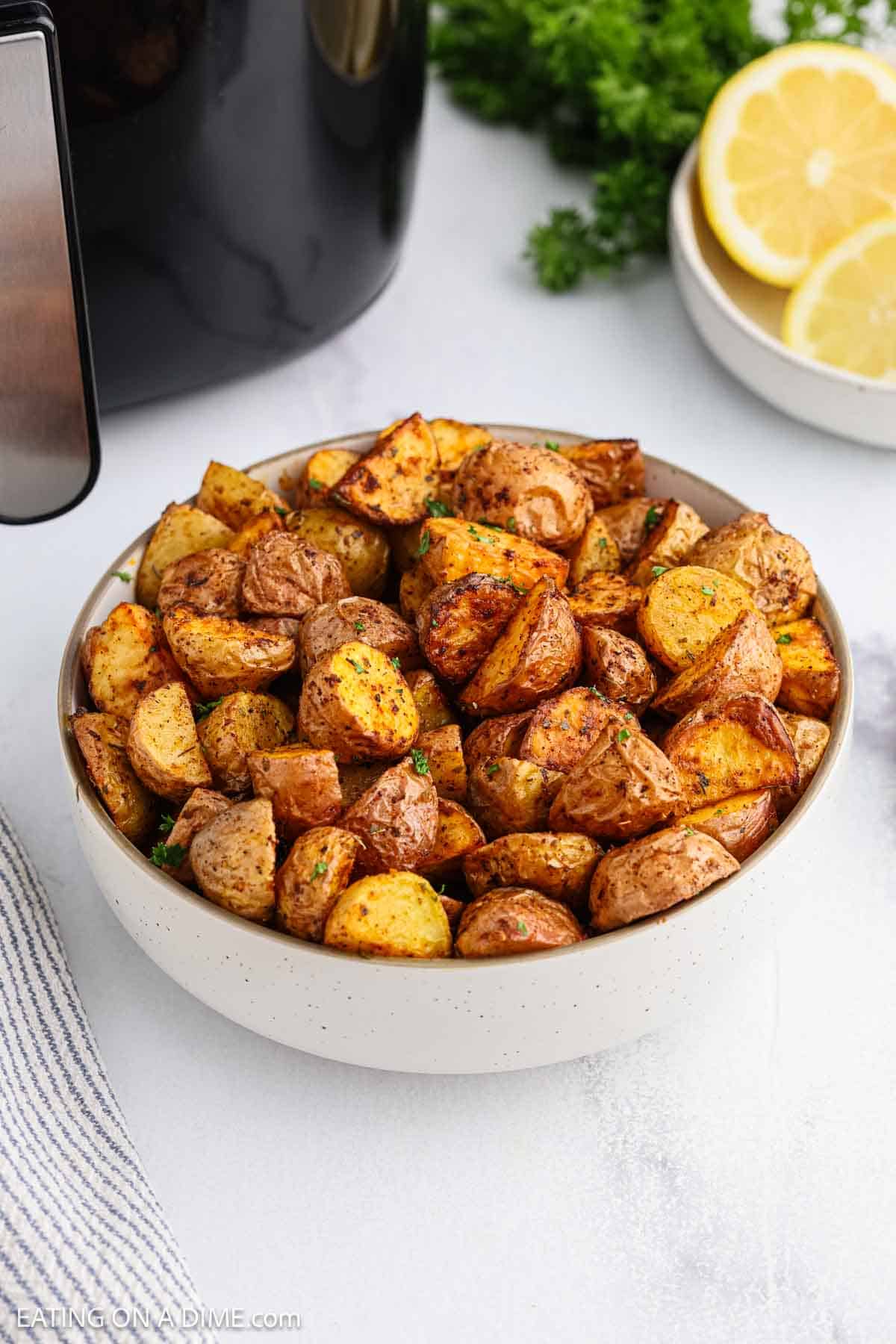 A bowl of crispy, golden-brown air fryer potatoes sits on a white surface near fresh parsley and sliced lemons, showcasing perfectly seasoned roasted chunks.