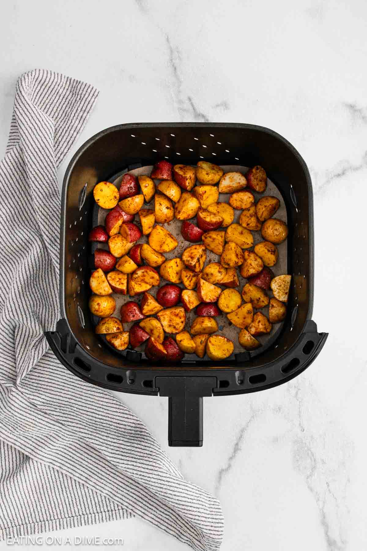 Air Fryer Potatoes seasoned and roasted to perfection in an air fryer basket, sitting on a white marble surface with a striped kitchen towel beside it.