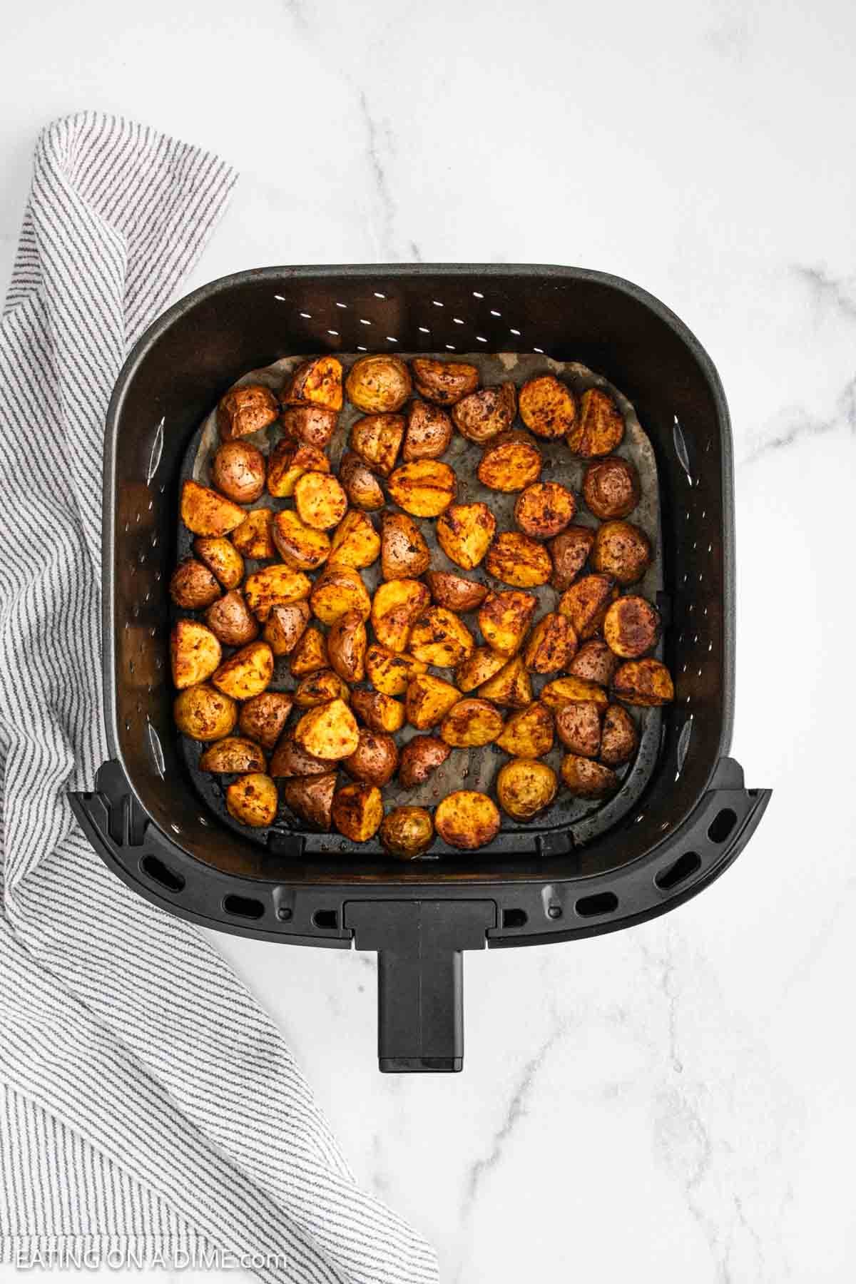 Overhead view of seasoned Air Fryer Potatoes in an air fryer basket, set on a white marble surface with a striped kitchen towel beside it.