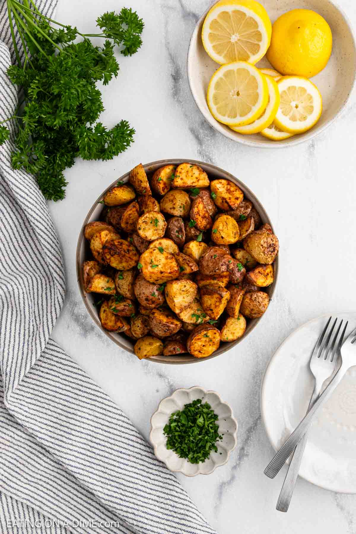 A bowl of crispy Air Fryer Potatoes garnished with herbs sits on a marble surface, surrounded by fresh parsley, sliced lemons, a small dish of chopped herbs, a striped towel, and two plates with forks.