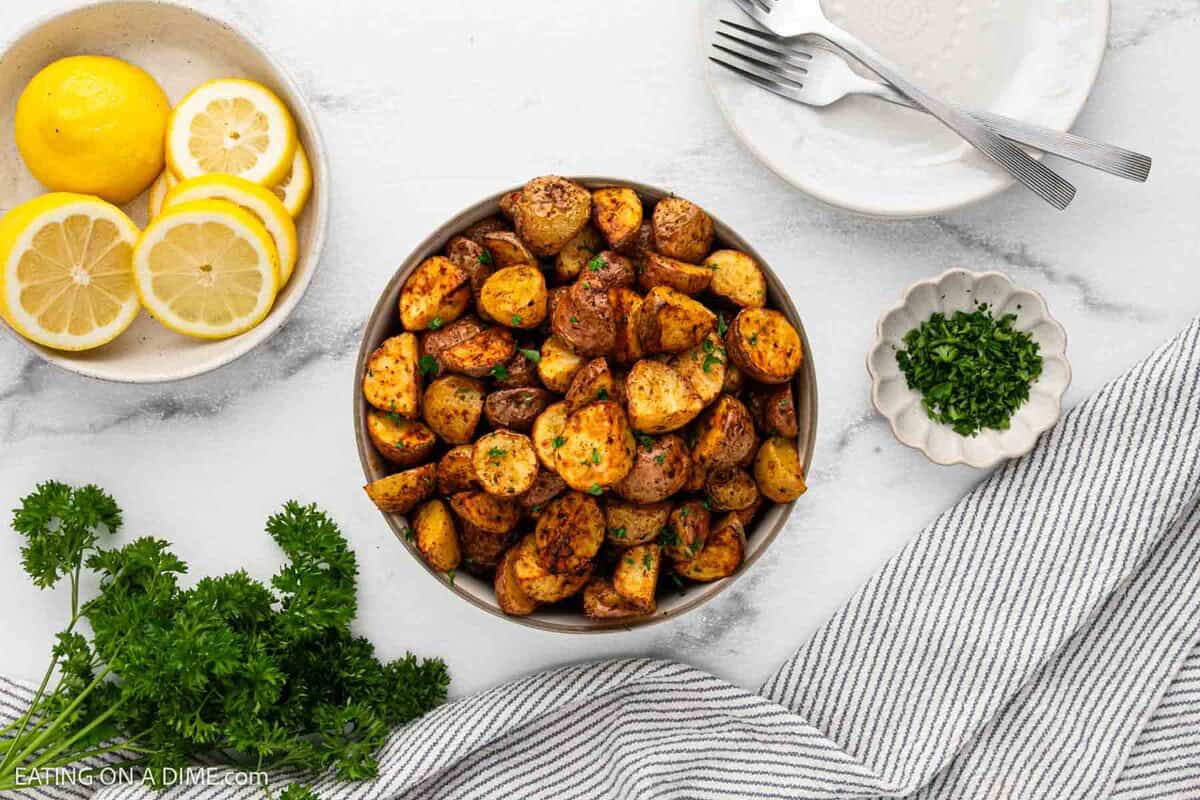 A bowl of crispy Air Fryer Potatoes is on a white surface, surrounded by fresh parsley, sliced lemons on a plate, a small dish of chopped herbs, a striped kitchen towel, and stacked plates with forks.