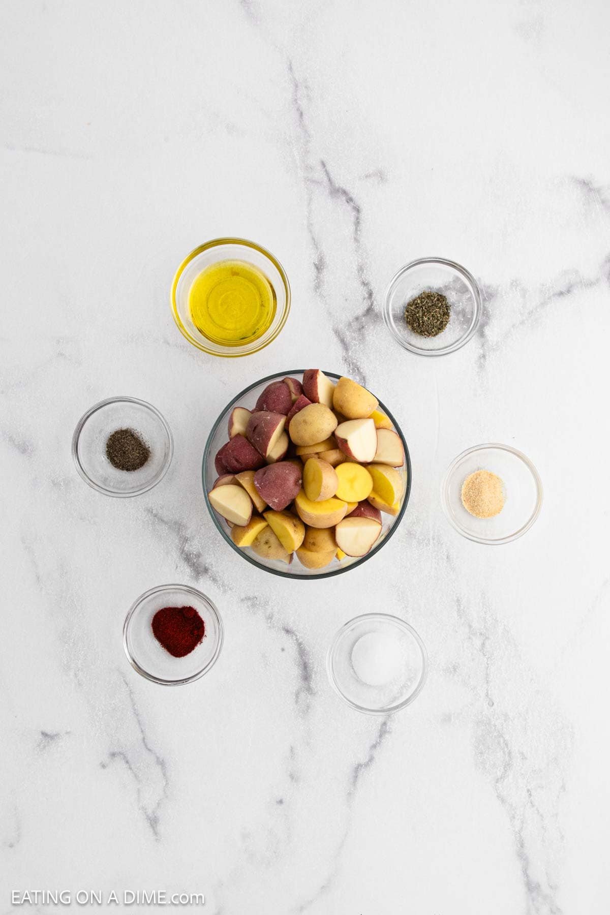 A bowl of quartered red and yellow baby potatoes is surrounded by small bowls of olive oil, salt, pepper, garlic powder, paprika, and Italian seasoningโperfect for creating delicious Air Fryer Potatoes on a white marble surface.