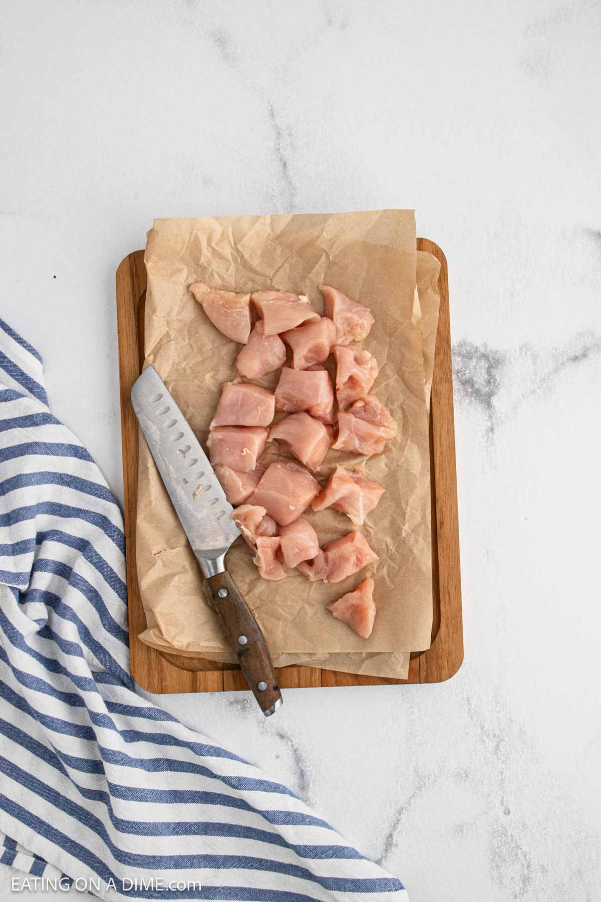 Raw chicken pieces, perfect for baked chicken bites, are arranged on parchment paper atop a wooden cutting board. A kitchen knife rests beside the chicken, and a blue-and-white striped towel is nearby on a light marble surface.