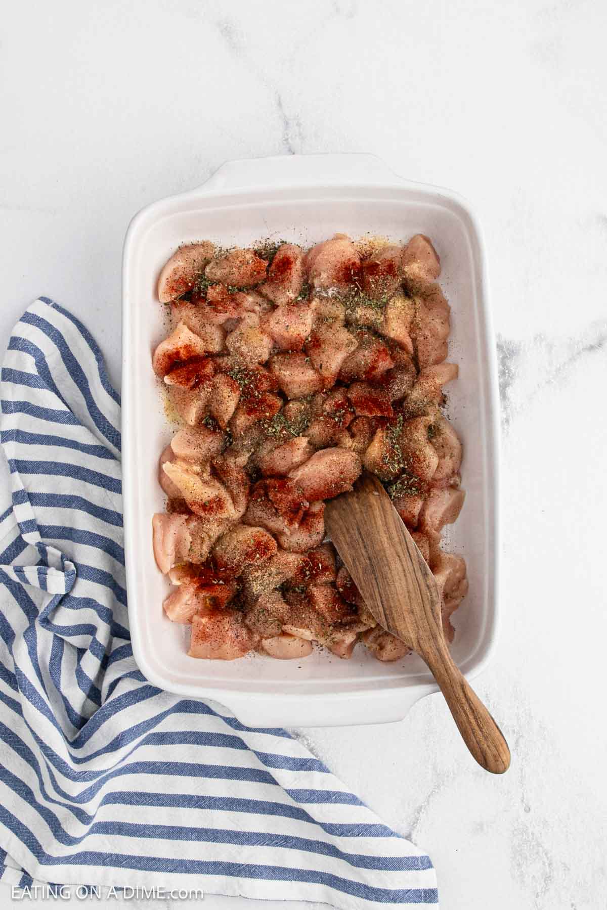 A white baking dish is filled with raw, seasoned Baked Chicken Bites, topped with spices. A wooden spoon rests in the dish, and a blue-and-white striped cloth lies beside it on a white marble surface.