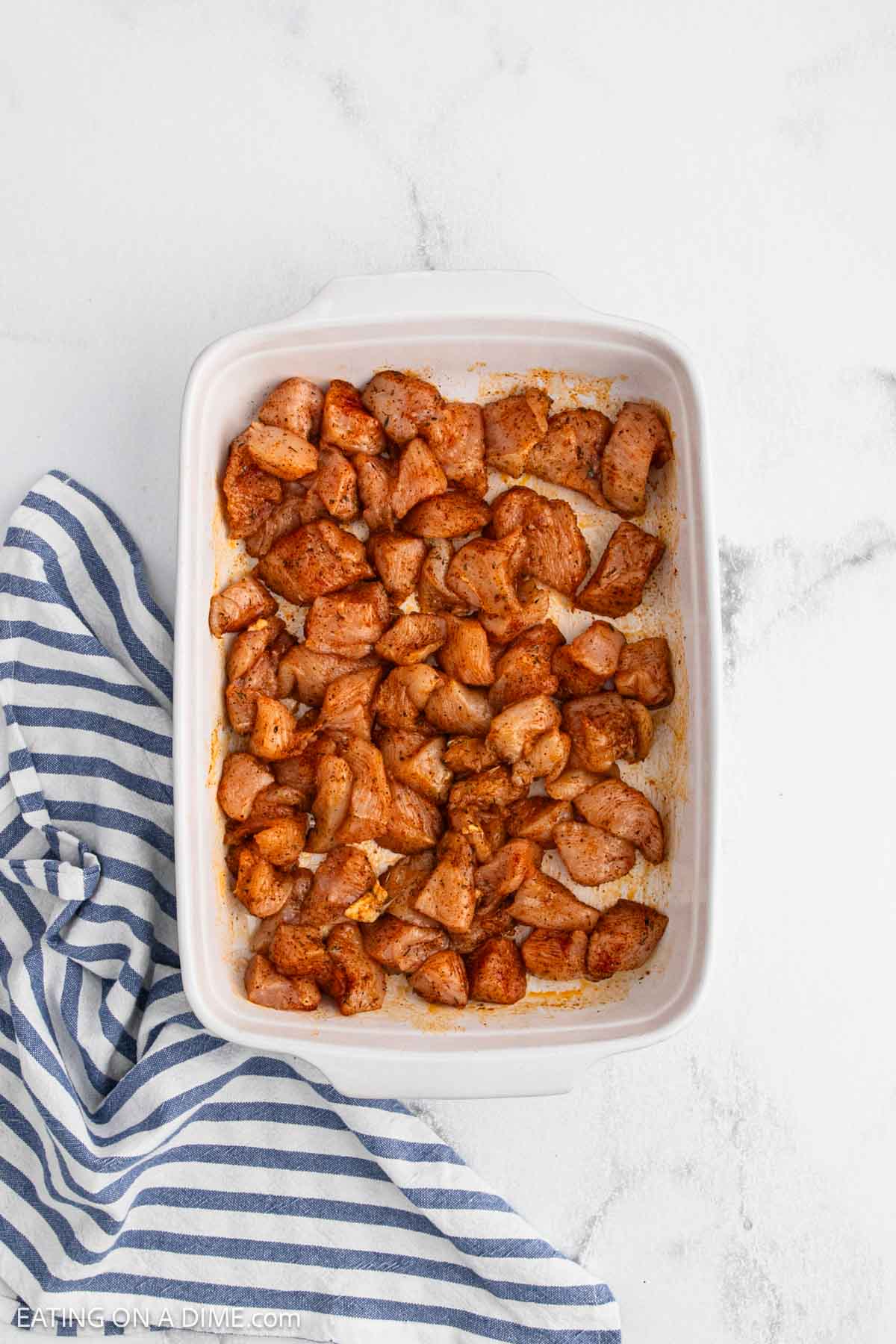 A white baking dish filled with Baked Chicken Bites sits on a white surface next to a blue and white striped kitchen towel, showcasing tender, seasoned chicken pieces ready to serve.