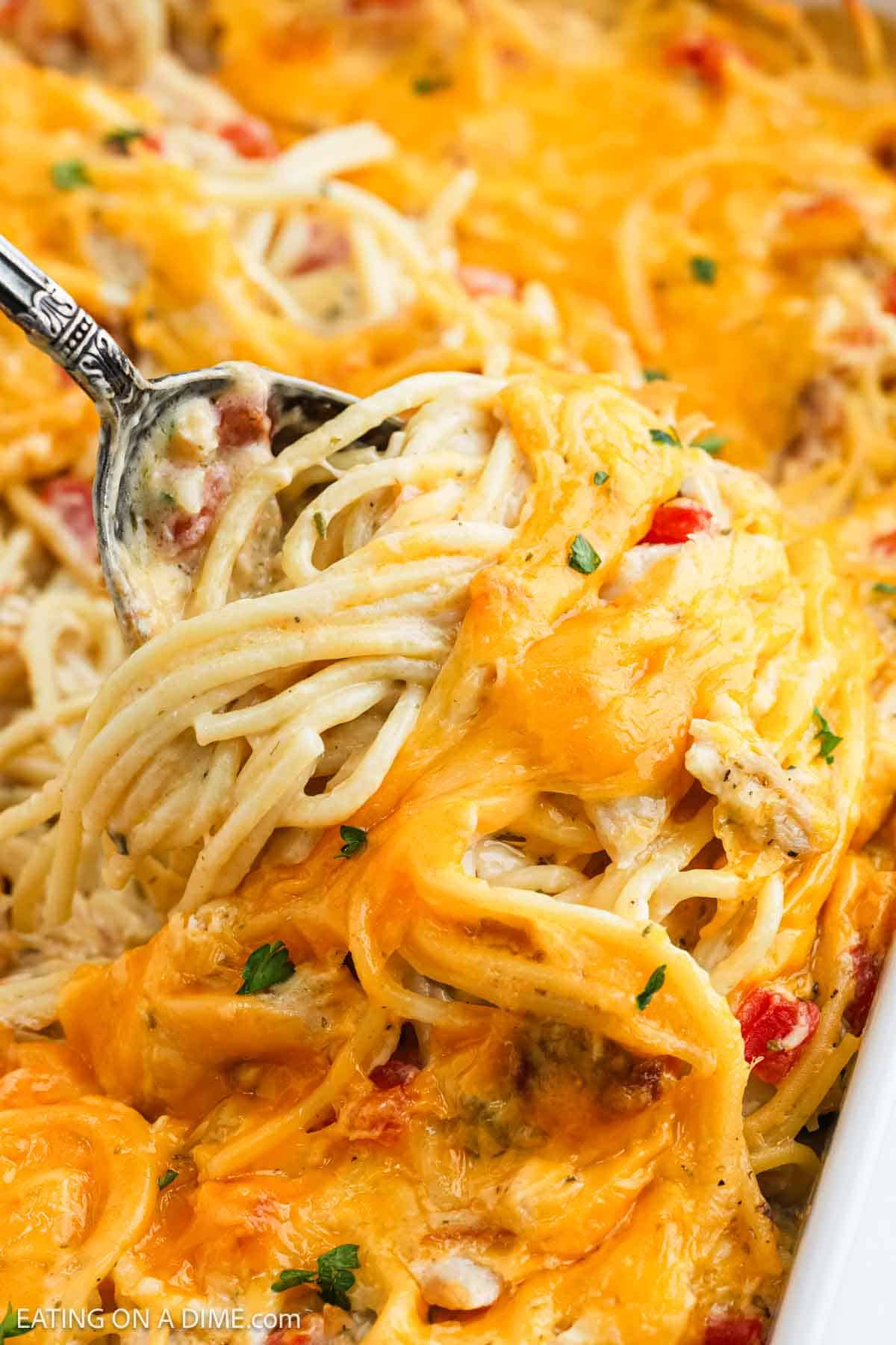 A close-up of creamy baked chicken spaghetti being scooped with a fork, topped with melted cheddar cheese and garnished with bits of parsley and red peppers.