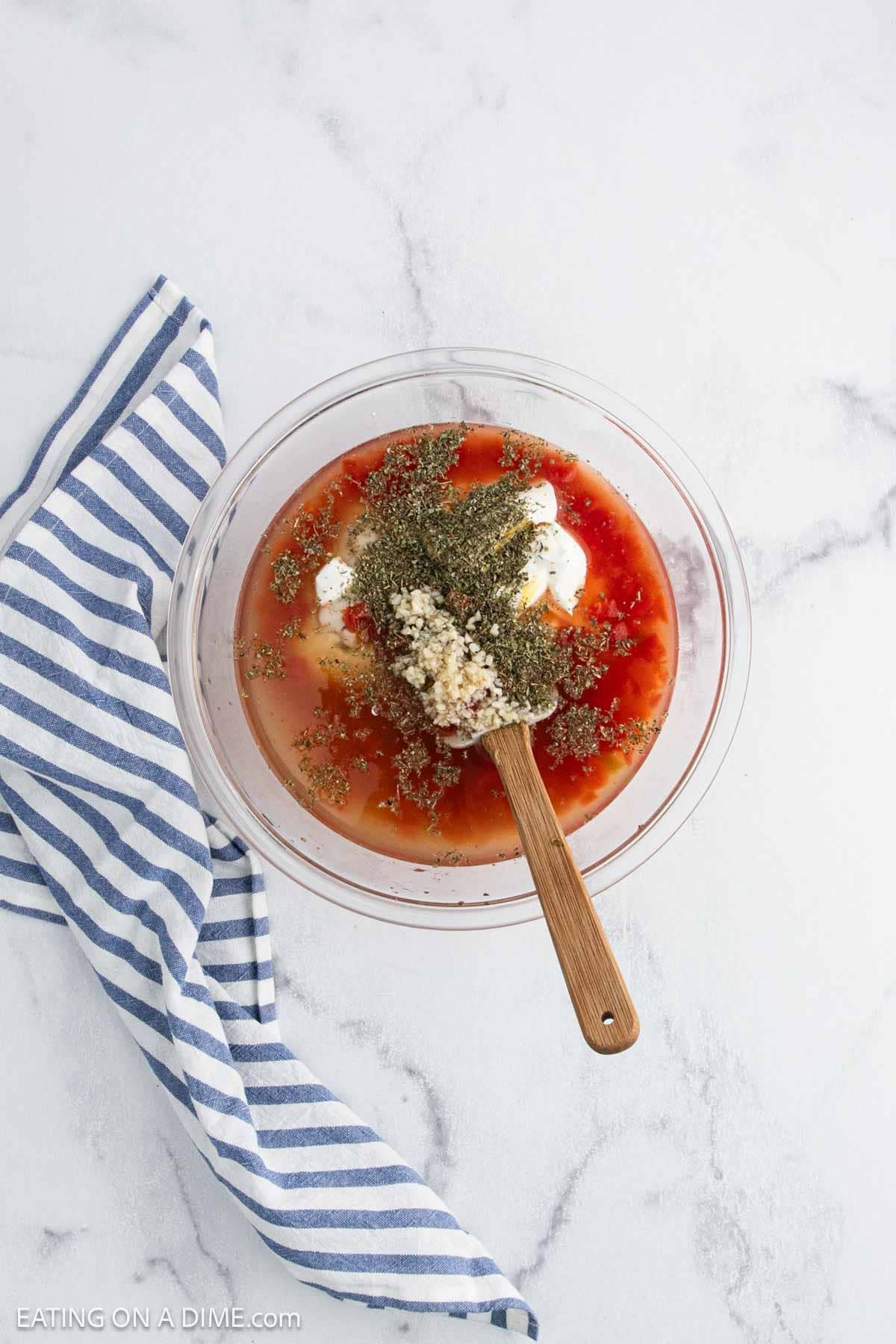 A glass bowl filled with red sauce, seasonings, minced garlic, and mayonnaiseโperfect for Baked Chicken Spaghettiโsits on a white marble surface next to a blue and white striped cloth. A wooden spoon rests in the bowl.