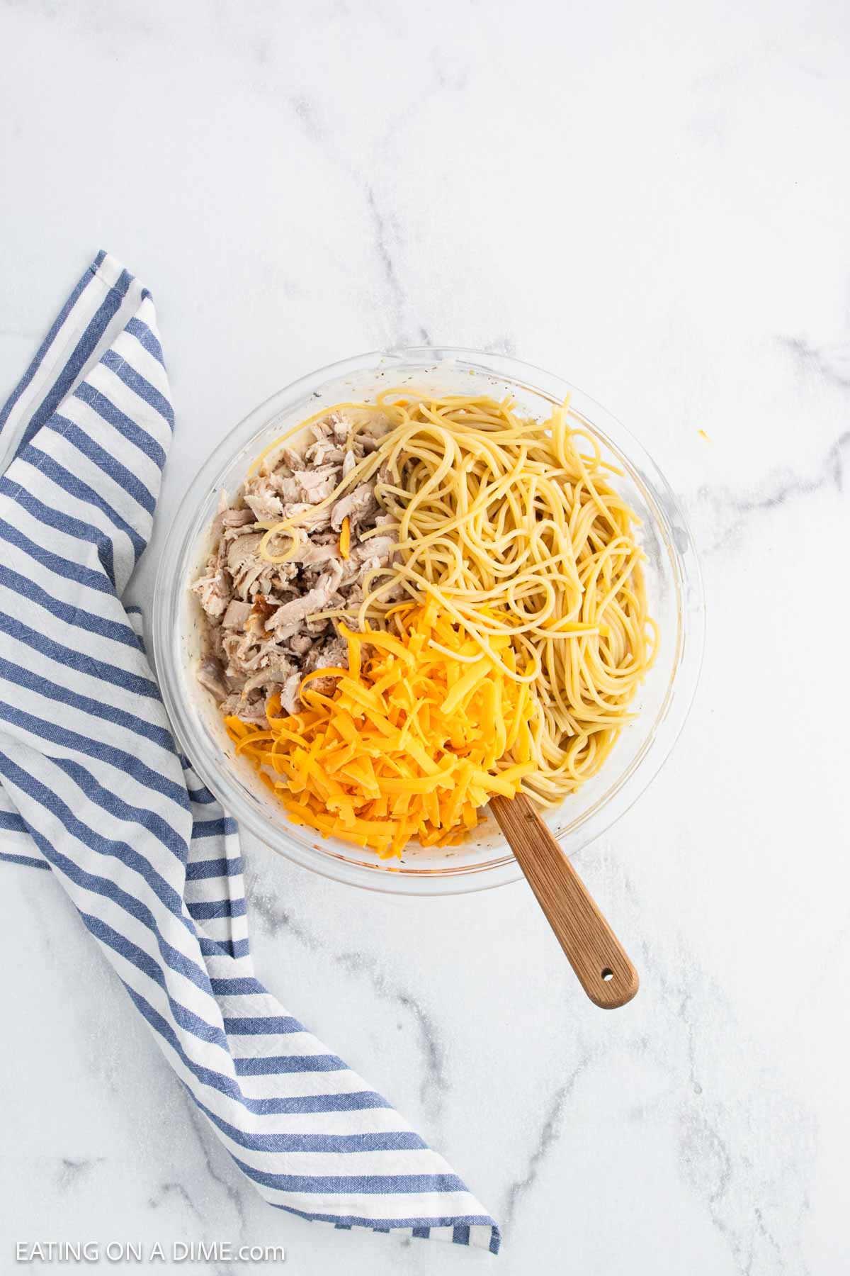 A glass bowl filled with baked chicken spaghettiโcooked spaghetti, shredded chicken, and cheddar cheeseโsits on a white marble surface beside a blue-and-white striped napkin. A wooden spoon rests inside the bowl.