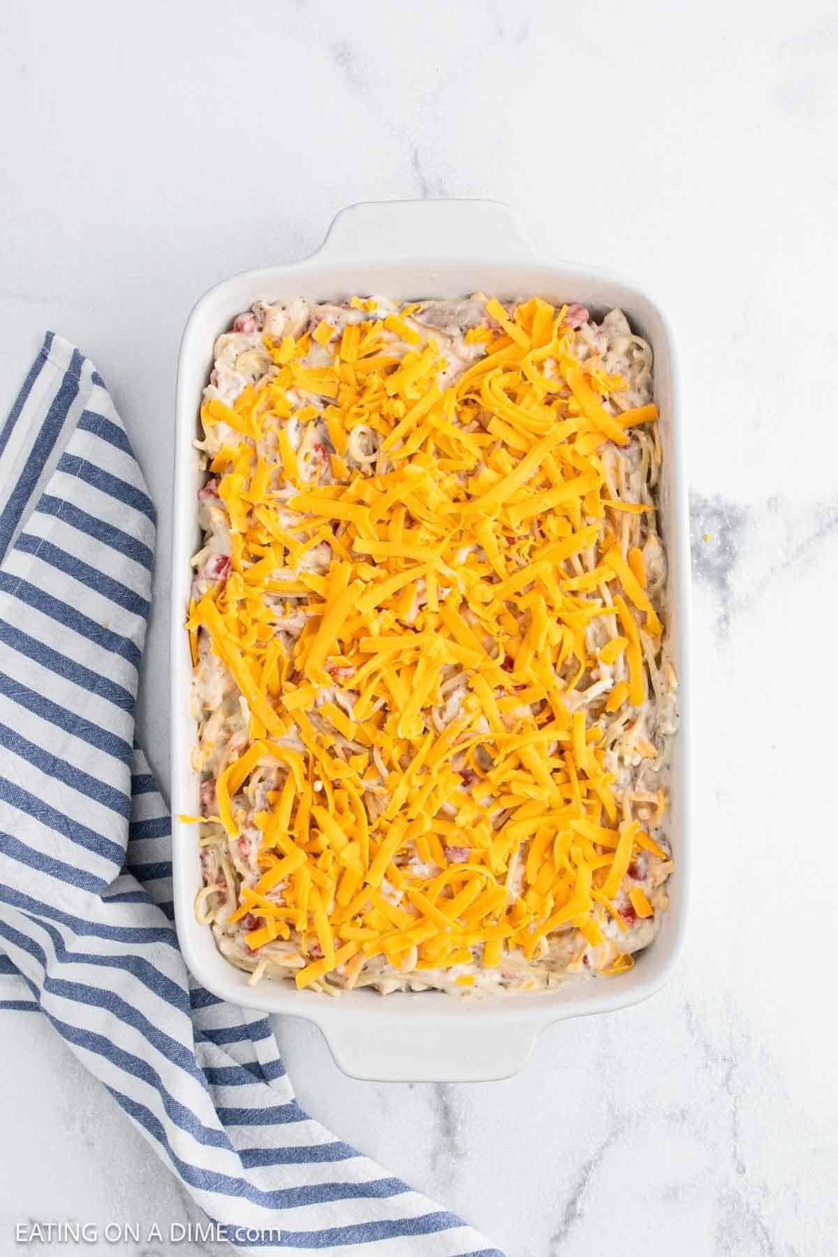 A white baking dish filled with creamy Baked Chicken Spaghetti, topped with a layer of shredded cheddar cheese, sits next to a blue and white striped kitchen towel on a light marble surface.