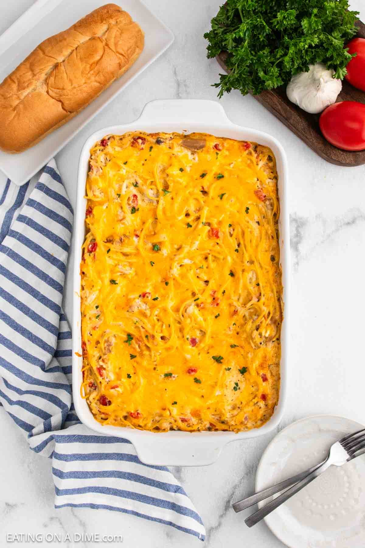 A baked Chicken Spaghetti casserole topped with melted cheddar cheese in a white dish, surrounded by a striped towel, sandwich bun, fresh parsley, garlic, tomatoes, and plates with forks.