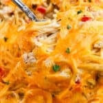 Close-up of a cheesy Baked Chicken Spaghetti dish with melted cheddar, small pieces of red bell pepper, and parsley garnish, with a fork lifting a portion.