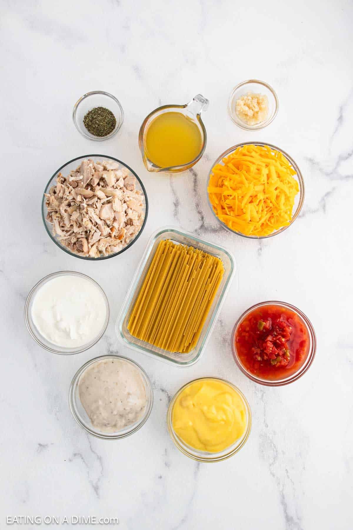 Ingredients for Baked Chicken Spaghetti arranged on a white surface, including dry spaghetti, shredded cheese, cooked chicken, canned soup, sour cream, diced tomatoes, broth, garlic, and Italian seasoning.