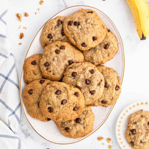 A white oval plate filled with homemade chocolate chip cookies sits on a marble surface, next to a striped cloth, a few cookie crumbs, and a ripe bananaโperfect inspiration for your next banana cookie recipe. A small plate holds an extra cookie.