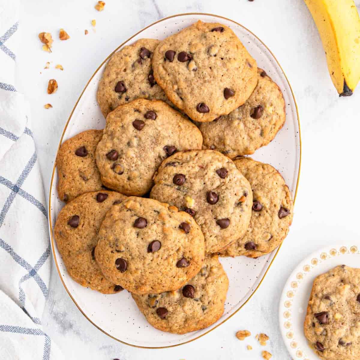 A white oval plate filled with homemade chocolate chip cookies sits on a marble surface, next to a striped cloth, a few cookie crumbs, and a ripe bananaโperfect inspiration for your next banana cookie recipe. A small plate holds an extra cookie.