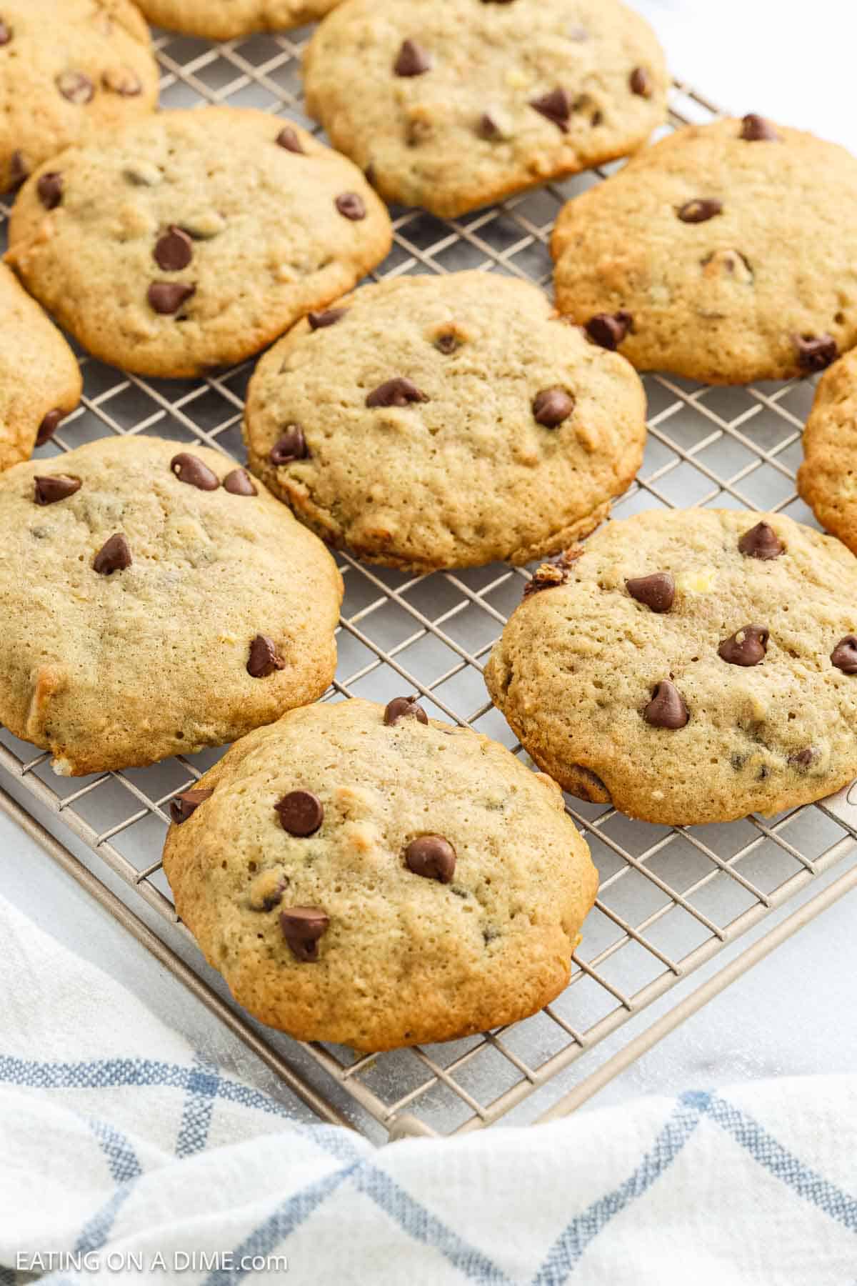 Freshly baked chocolate chip cookies cooling on a wire rack, with a blue and white striped cloth underneath. These golden brown treats use our Banana Cookie Recipe for an irresistible twist and extra soft, flavorful bites.