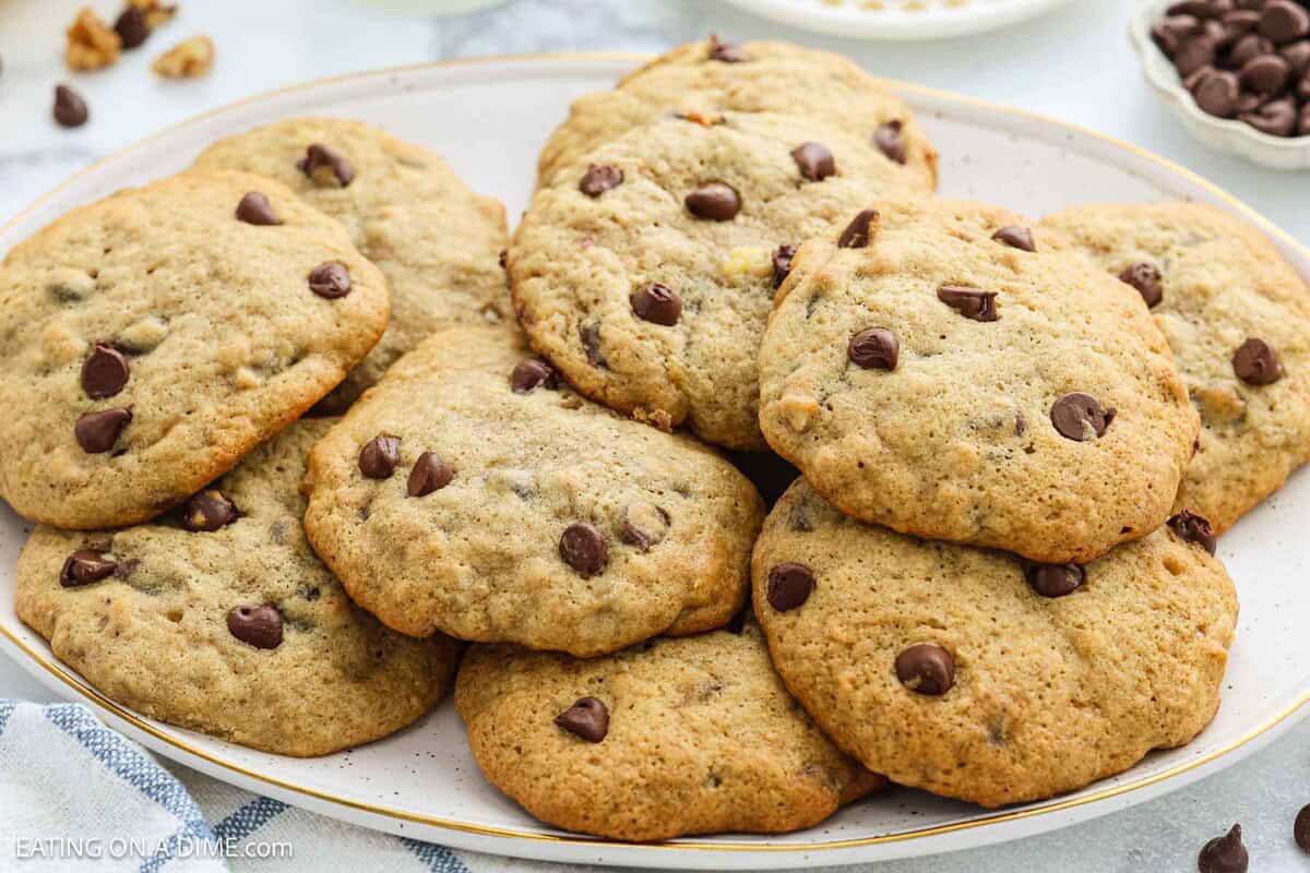 A plate piled with homemade banana cookies, golden brown and studded with chocolate chips, sits on a white surface with more cookies and ingredients from the tasty banana cookie recipe visible in the background.