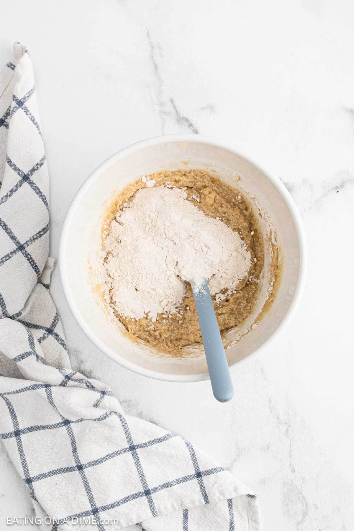 A white mixing bowl with partially mixed batter for a Banana Cookie Recipe sits on a white marble surface. A blue-handled spatula is in the bowl, and a blue-and-white checkered cloth is nearby.