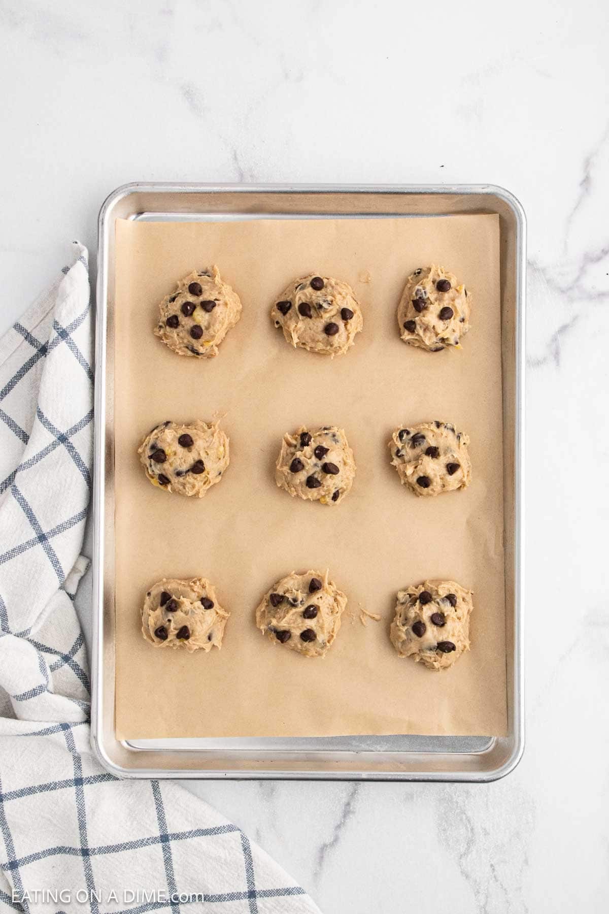 A metal baking sheet lined with parchment paper holds nine scoops of banana cookie dough, ready to bake. A white and blue plaid kitchen towel is beside the tray on a marble countertop, perfect for trying out a new banana cookie recipe.