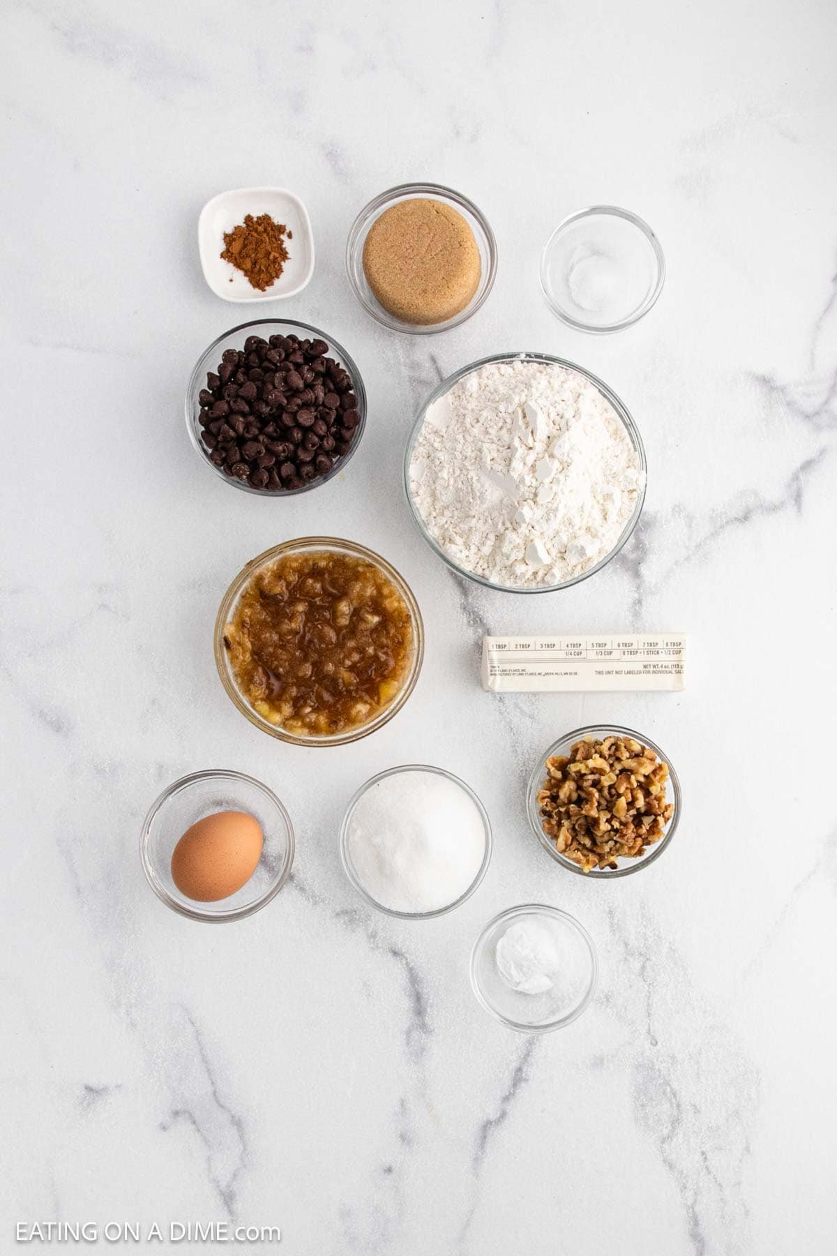 Overhead view of baking ingredients in small bowls on a white surface, perfect for a Banana Cookie Recipe—included are flour, chocolate chips, brown sugar, walnuts, egg, butter, applesauce, spices, and baking powder.