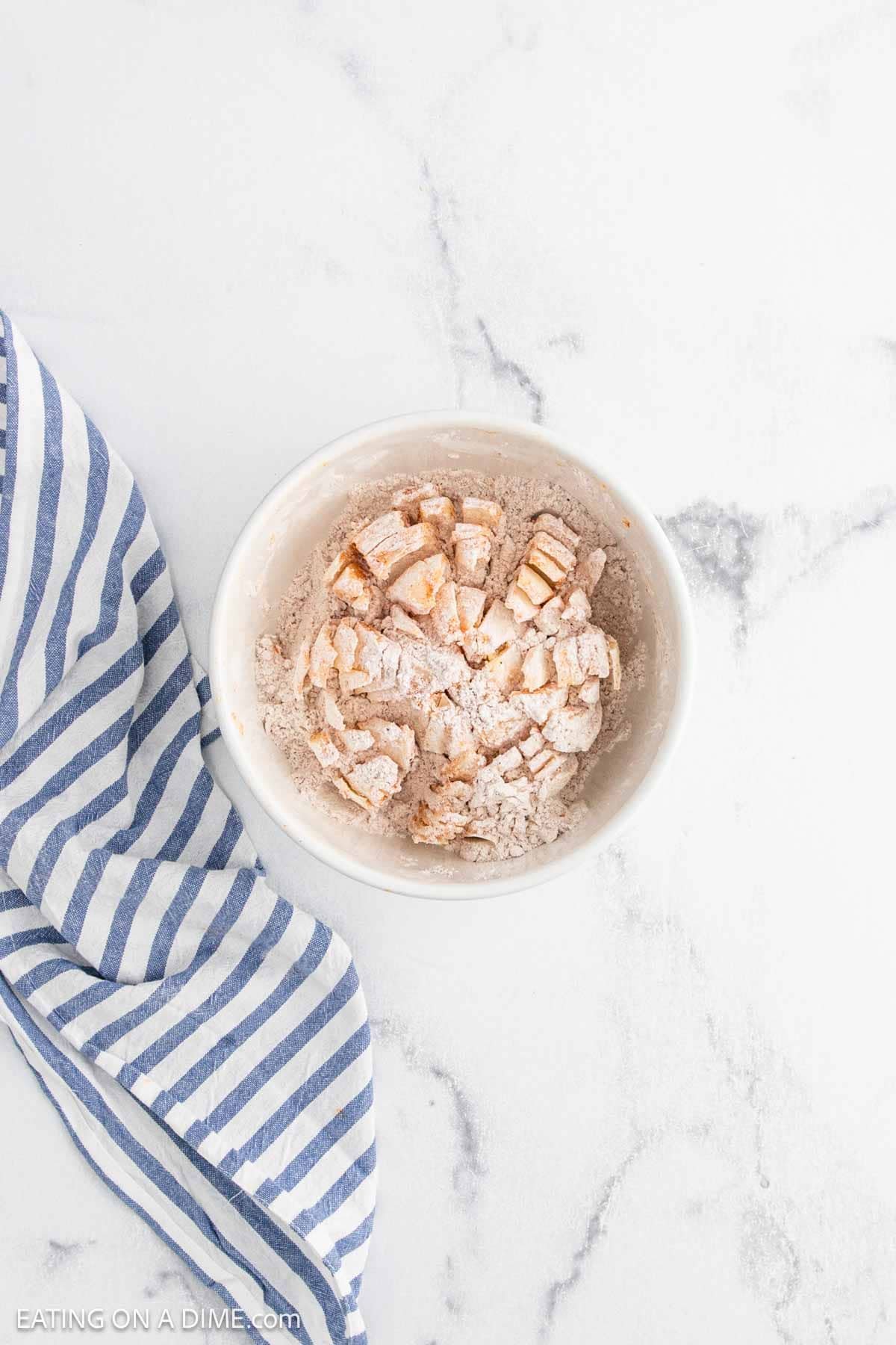 A white bowl filled with diced apples coated in flour and cinnamon sits on a marble surface next to a blue and white striped kitchen towel, ready for baking—reminiscent of prepping a Bloomin Onion for the oven.
