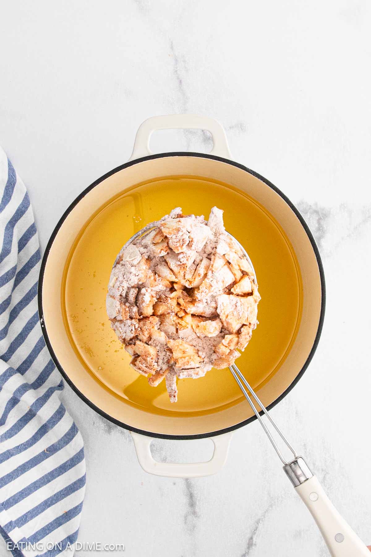 A battered Bloomin Onion is being lowered into a pot of hot oil with a slotted spatula, next to a blue and white striped towel on a white surface.