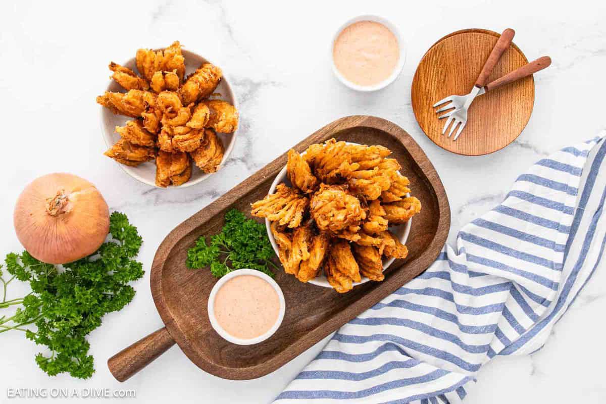 A wooden board with a bowl of crispy fried onion petals, a dish of dipping sauce, fresh parsley, an onion, and a bowl of more Bloomin Onion appetizer petals rest beside mini forks and a striped napkin on a marble surface.