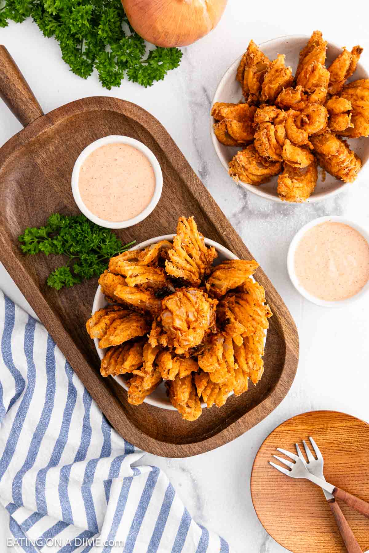 A wooden serving board features a crispy Bloomin Onion with dipping sauce, garnished with parsley. Nearby, two small bowls hold fried onion petals and extra sauce, next to a striped napkin and a plate with a fork.