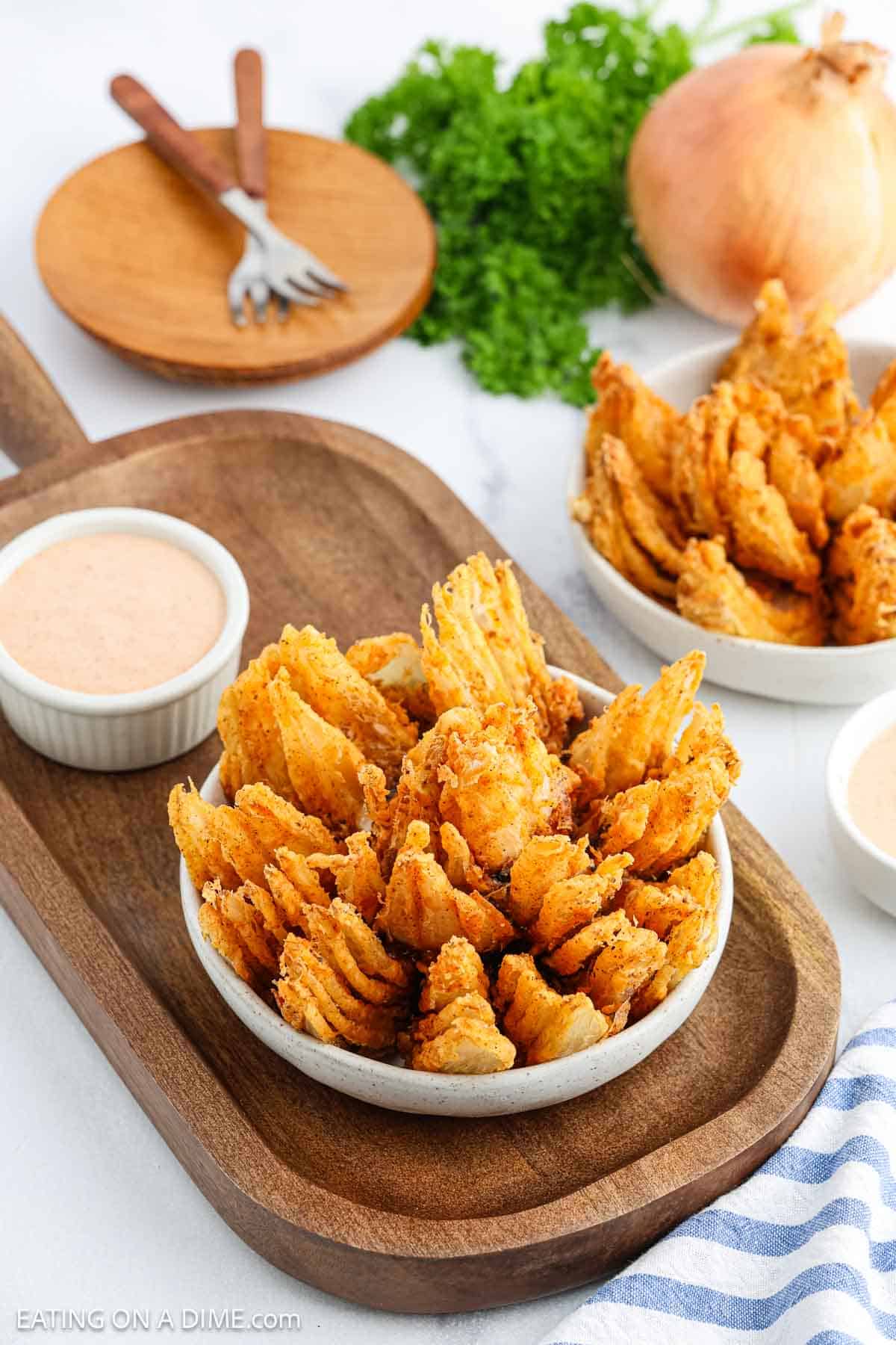 A bowl of Bloomin Onion petals served with a side of dipping sauce on a wooden tray. In the background, another plate of onion petals, fresh parsley, and a whole onion complete the scene.