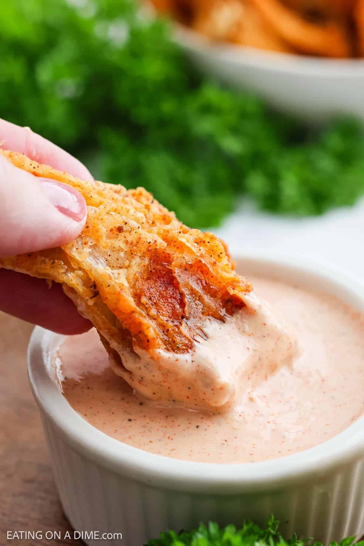 A hand dips a crispy pepperoni pizza chip into a creamy, seasoned dipping sauce in a small white ramekin, reminiscent of Bloomin Onion sauce, with fresh parsley and more chips visible in the background.