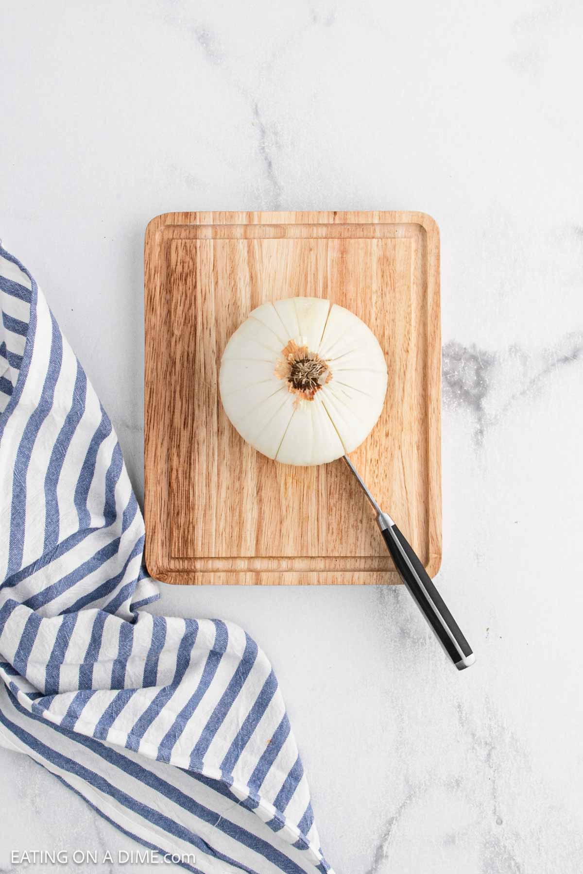 A whole peeled onion with the root end intact sits on a wooden cutting board, ready to be transformed into a Bloomin Onion appetizer like those at Outback Steakhouse. A knife is partially inserted, with a blue and white striped cloth nearby on a light surface.