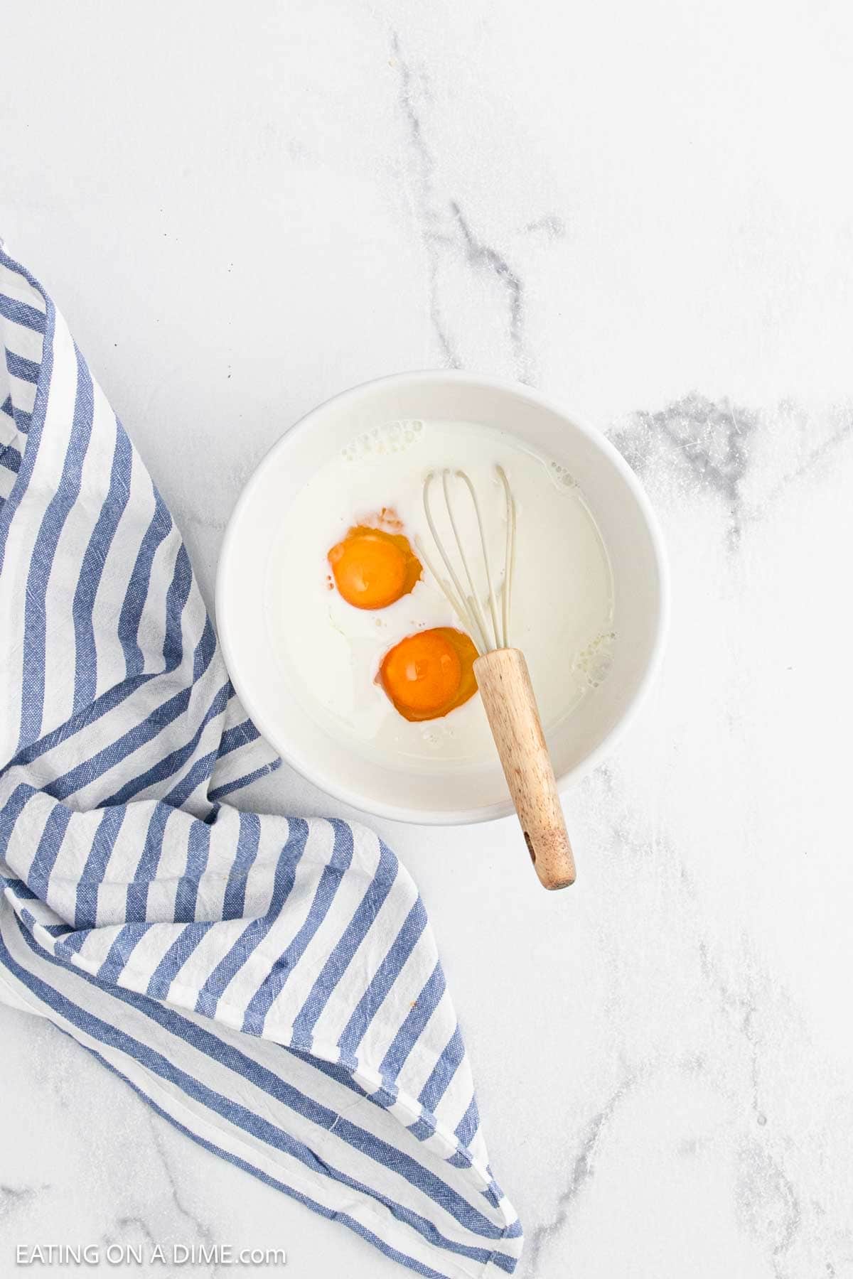 A white bowl with two eggs, milk, and a small whisk sits on a white surface next to a blue and white striped cloth—perfect for prepping Bloomin Onion batter.
