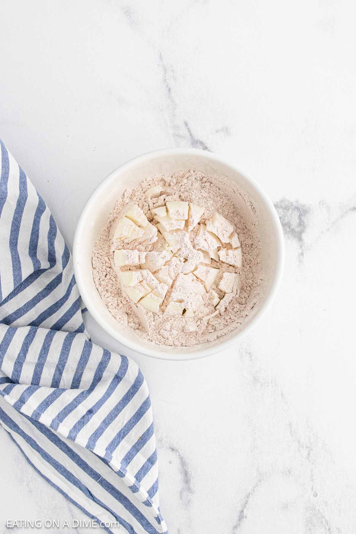 A white bowl filled with flour and cubed butter, perfect for crafting a Bloomin Onion batter, sits on a white marble surface next to a blue and white striped cloth.