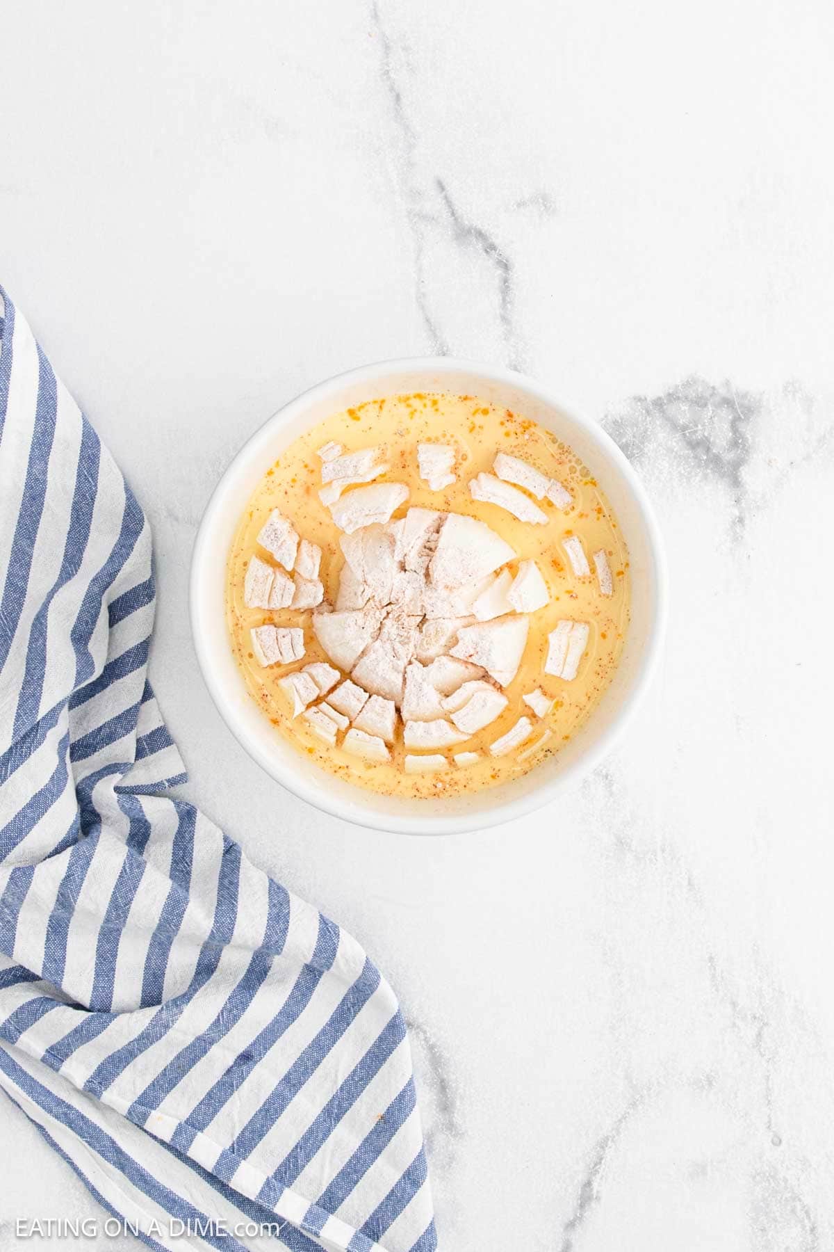 A bowl of banana pudding topped with sliced bananas and crumbled vanilla wafers sits next to a blue and white striped cloth on a white marble surface, capturing the comfort of homemade treats—no Bloomin Onion needed.