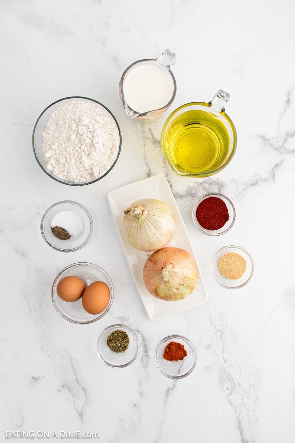 Ingredients for a recipe arranged on a white surface, featuring two whole onions ready to become a Bloomin Onion, along with eggs, flour, oil, milk, and small bowls of paprika, black pepper, garlic powder, and dried herbs.