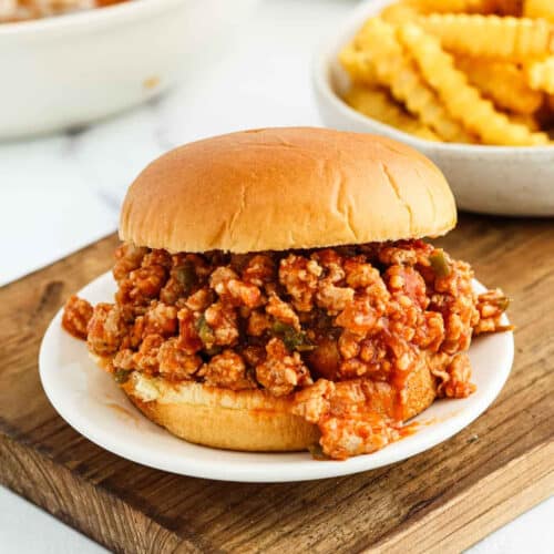 A Cajun Chicken Sloppy Joes sandwich filled with seasoned ground meat and zesty sauce sits on a white plate, with a bowl of crinkle-cut French fries in the background.