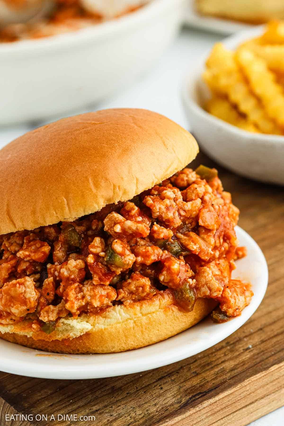 A Chicken Sloppy Joes sandwich filled with a flavorful, saucy ground chicken mixture sits on a white plate. In the background, there is a bowl of crinkle-cut French fries.