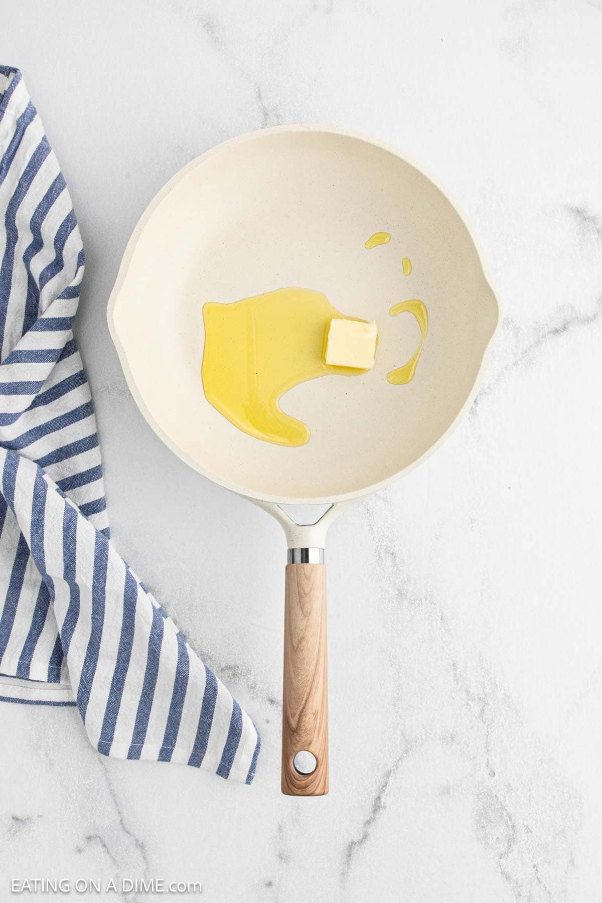A white frying pan with a wooden handle contains a pat of butter and melted butter, ready for making Cajun chicken or other delicious Cajun recipes. The pan sits on a white marble surface next to a blue and white striped kitchen towel.
