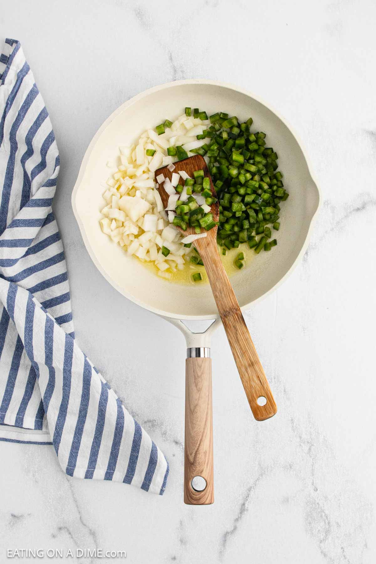 A white skillet with chopped green bell peppers and onions for Cajun Chicken Sloppy Joes is being stirred with a wooden spatula, sitting on a white marble surface next to a blue and white striped kitchen towel.