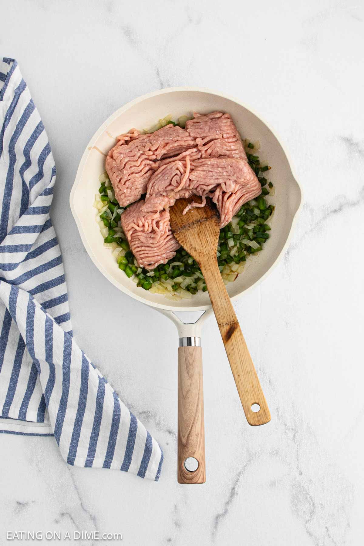 A white skillet with raw ground turkey, chopped onions, and green peppers sits on a marble surface—perfect for making Cajun Chicken Sloppy Joes. A wooden spoon rests in the pan, and a blue-and-white striped towel is nearby.