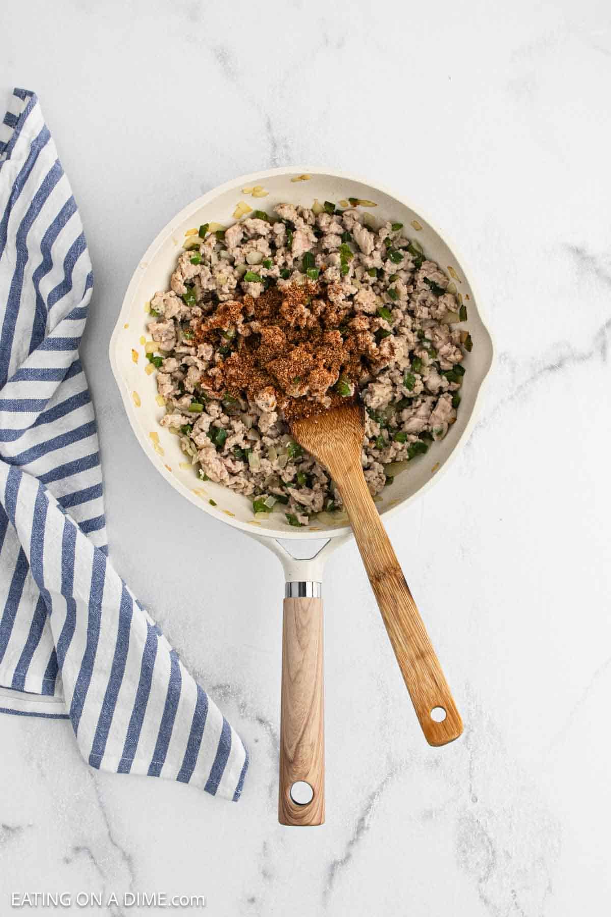 A white skillet filled with cooked ground meat, chopped green peppers, and spices for Cajun Chicken Sloppy Joes is being stirred with a wooden spoon, next to a blue and white striped kitchen towel on a marble surface.