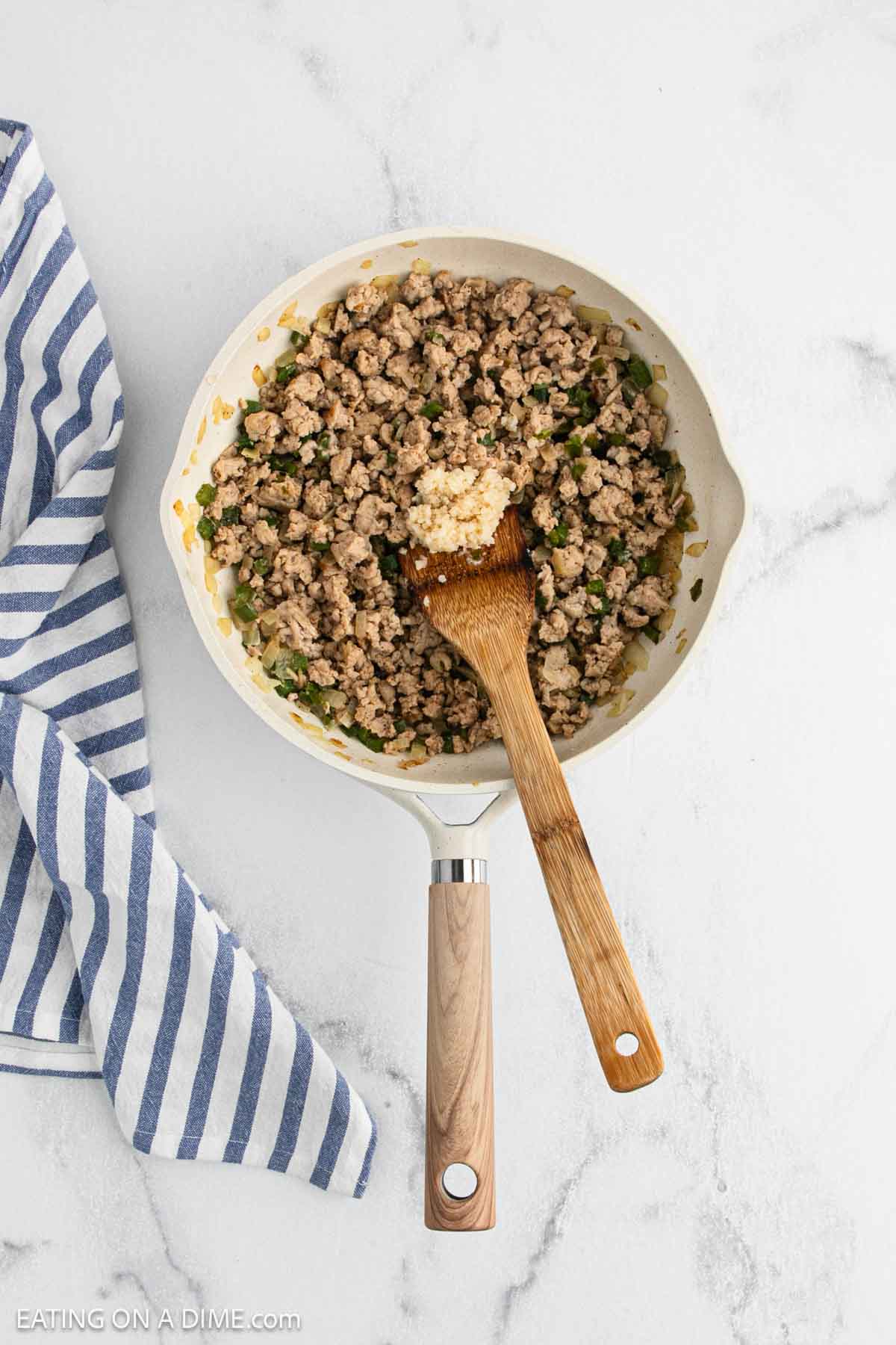 A skillet filled with cooked Cajun Chicken and chopped vegetables sits on a white surface. A wooden spoon rests in the pan, hinting at flavorful Cajun Chicken Sloppy Joes, while a blue-and-white striped cloth is nearby.