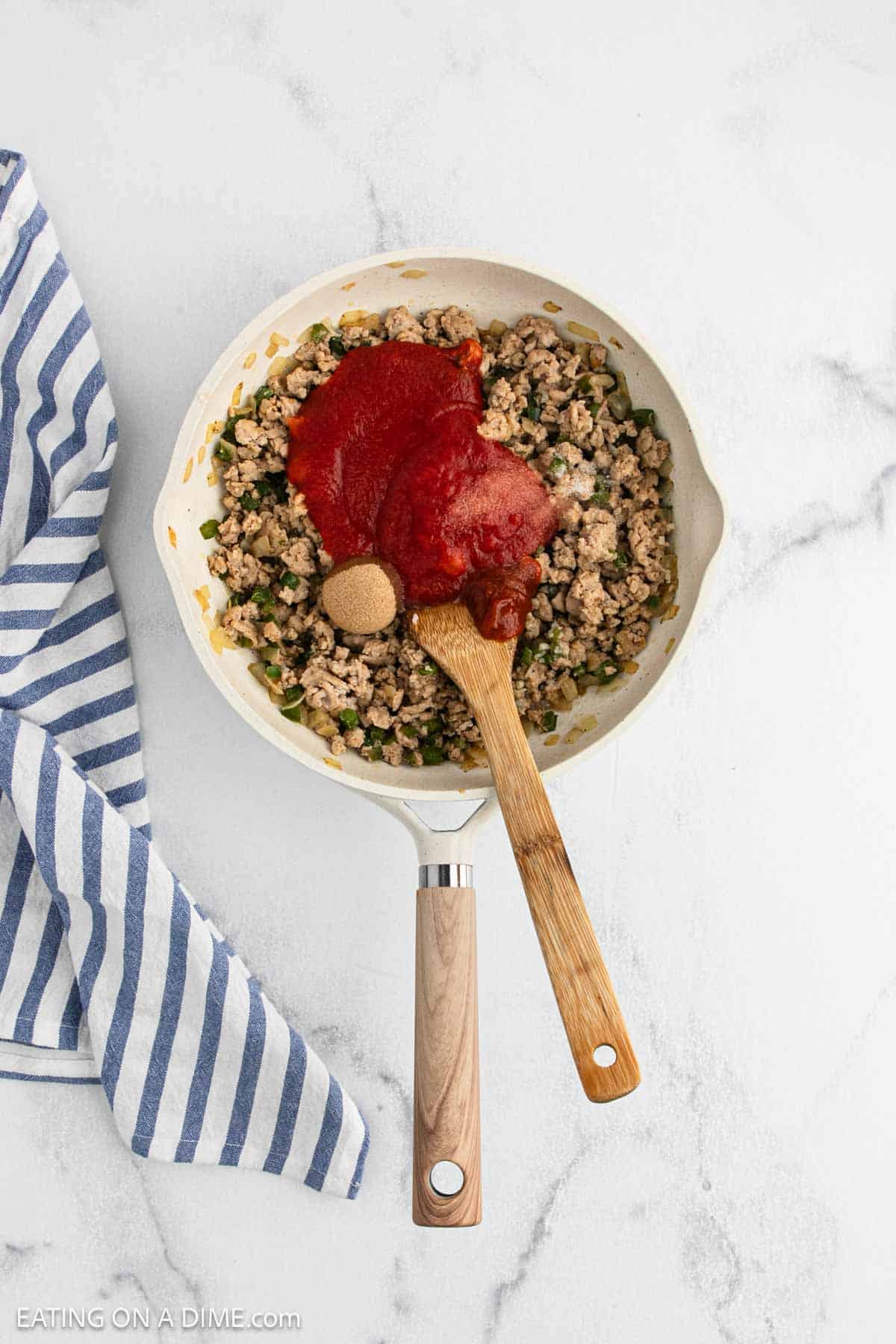 A white skillet with cooked ground meat for Sloppy Joes, topped with tomato sauce and brown sugar, sits on a marble surface next to a blue and white striped kitchen towel. A wooden spoon rests in the skillet.