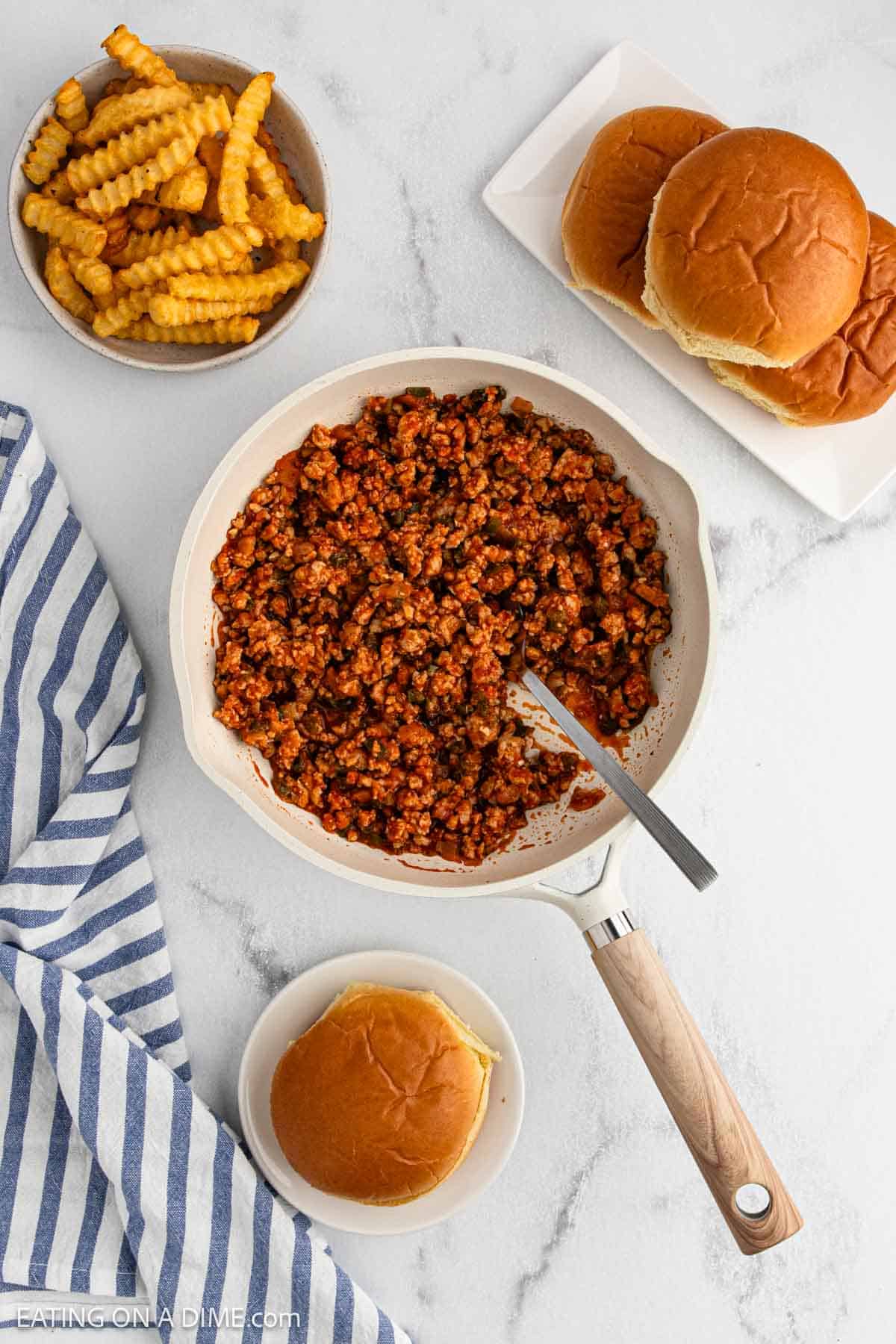 A skillet of Cajun Chicken Sloppy Joes sits on a white surface next to crinkle-cut fries, a stack of hamburger buns, and a single assembled sandwich. A blue-striped towel lies nearby.