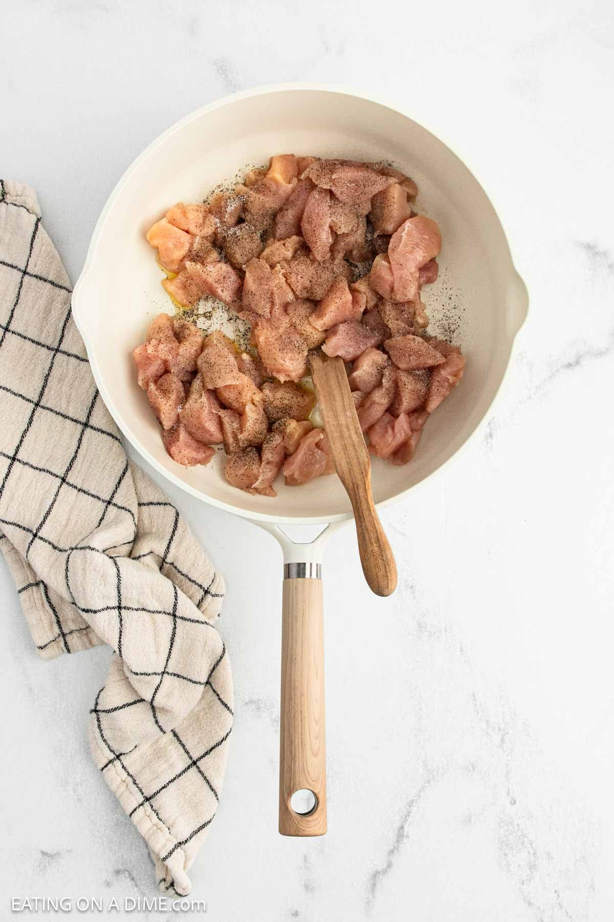 A white skillet with raw, seasoned Cajun Chicken pieces and a wooden spatula on a marble surface, next to a beige kitchen towel with a black grid pattern.
