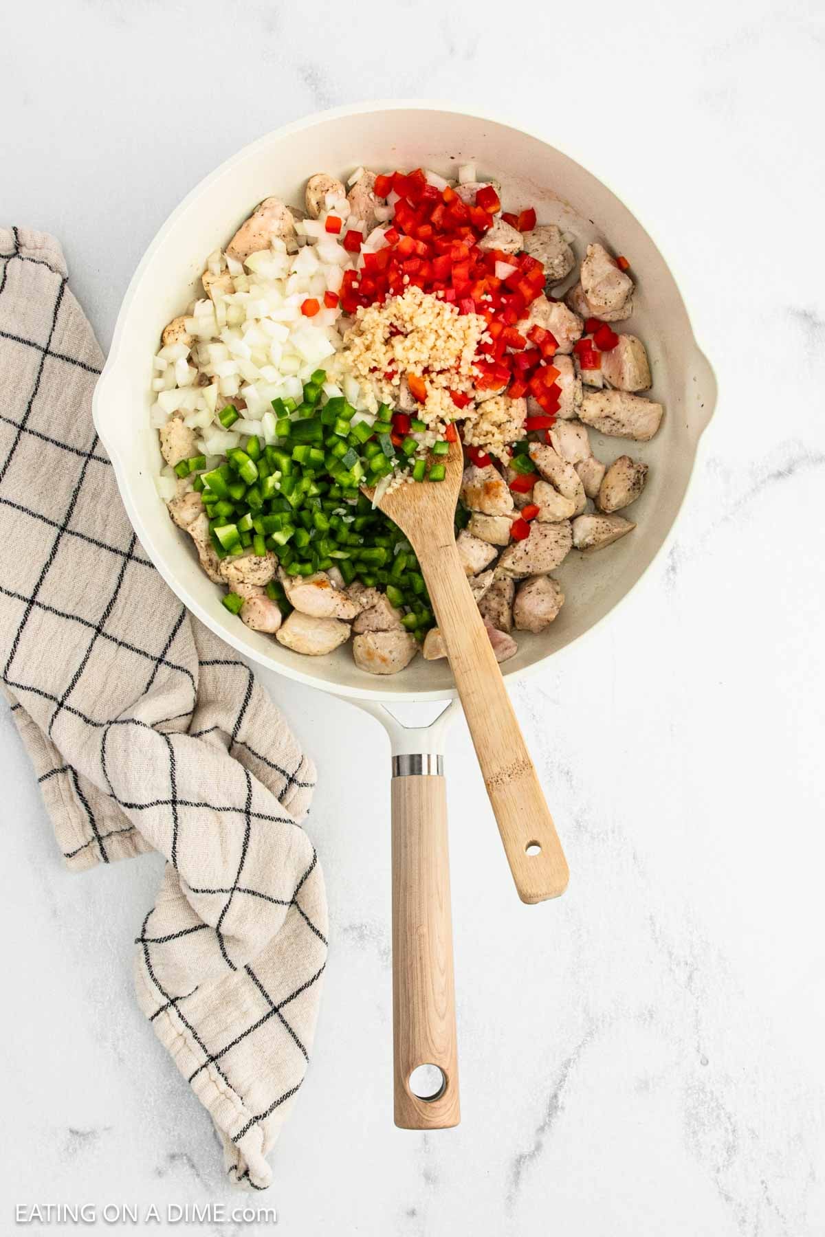 A skillet filled with cooked diced Cajun Chicken, chopped onions, red and green bell peppers, and minced garlic, with a wooden spoon resting inside. A checkered kitchen towel lies beside the skillet on a white surface.