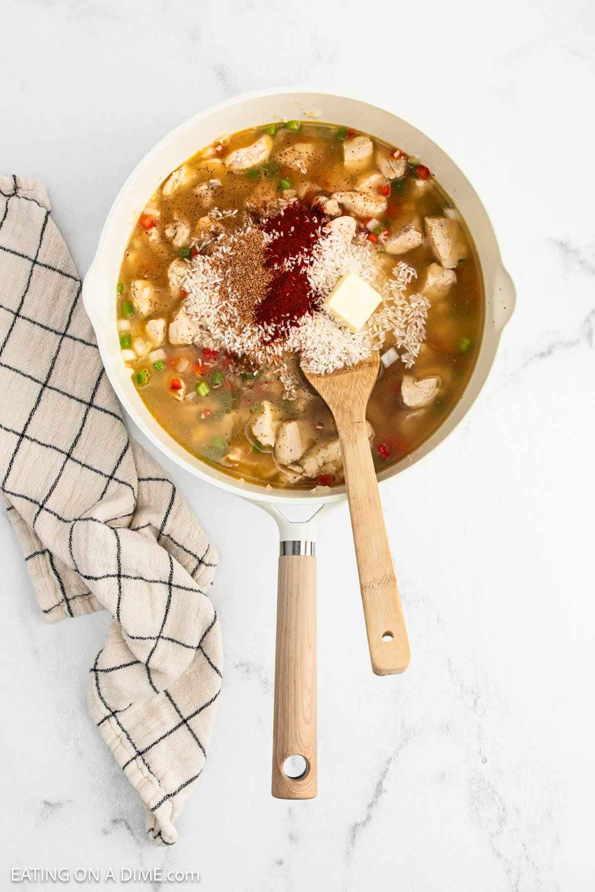 A skillet on a marble countertop contains Cajun Chicken and Rice with broth, spices, butter, and chopped vegetables. A wooden spoon rests inside the skillet, and a folded checkered towel is beside it.