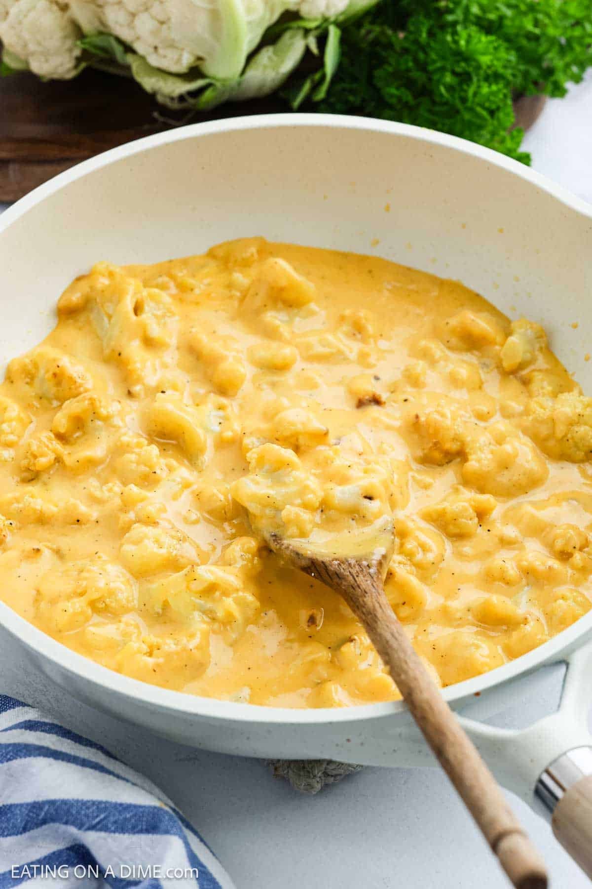 A creamy Cauliflower Mac and Cheese mixture is being stirred in a white pan with a wooden spoon. Fresh cauliflower and leafy greens can be seen in the background.