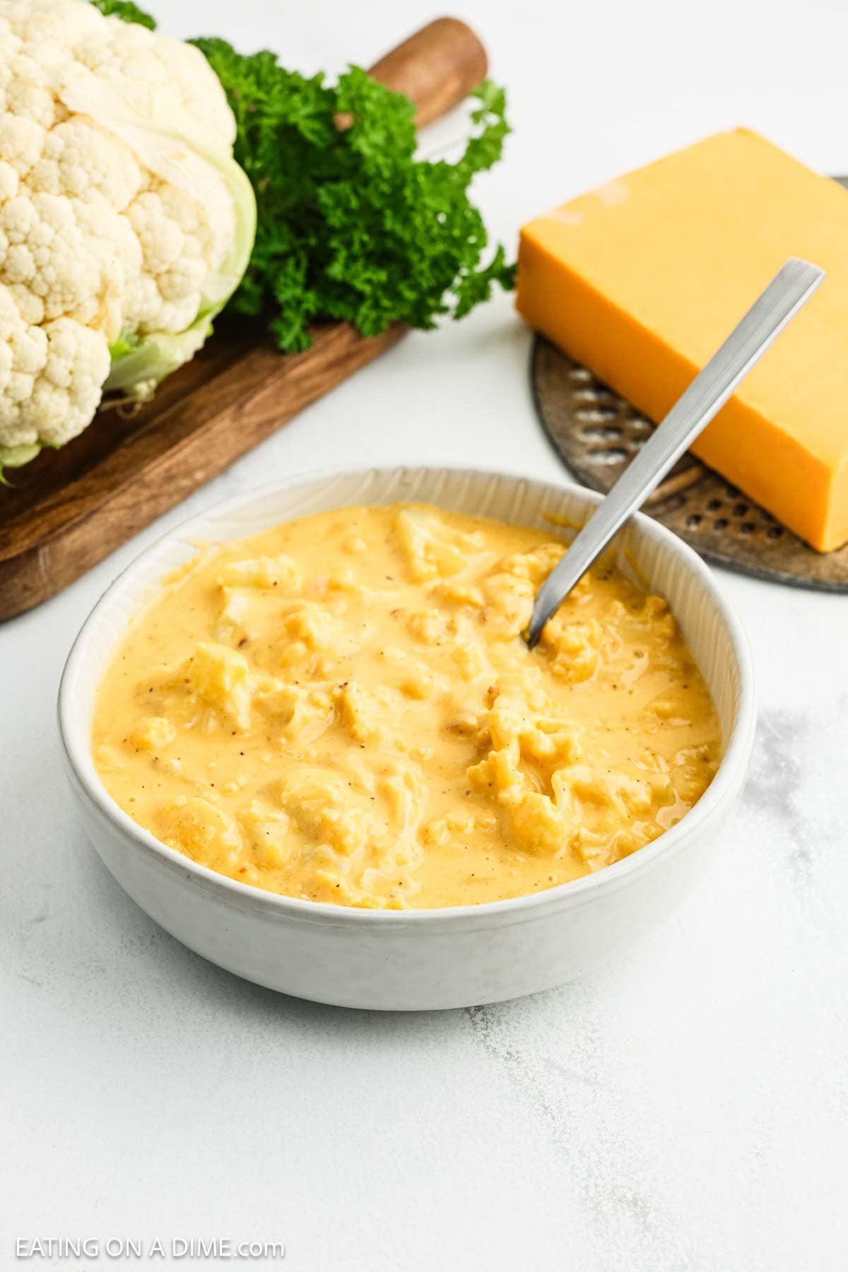 A bowl of creamy cauliflower cheese soup with a spoon, next to a block of cheddar cheese and a head of cauliflower on a cutting boardโgarnished with parsley and inspired by classic mac and cheese flavors.