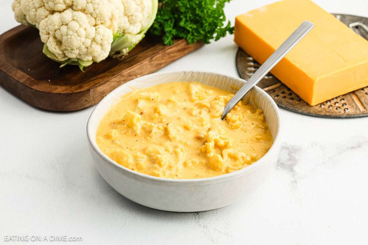 A bowl of creamy cauliflower cheese soup with a spoon, placed on a white surface. Behind it are a block of cheddar cheese, a head of cauliflower, and some fresh parsleyโperfect for fans of Cauliflower Mac and Cheese.
